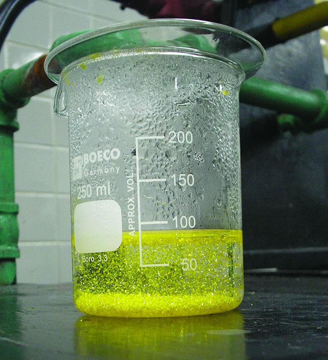 A photograph is shown of a yellow green opaque substance swirled through a clear, colorless liquid in a test tube.